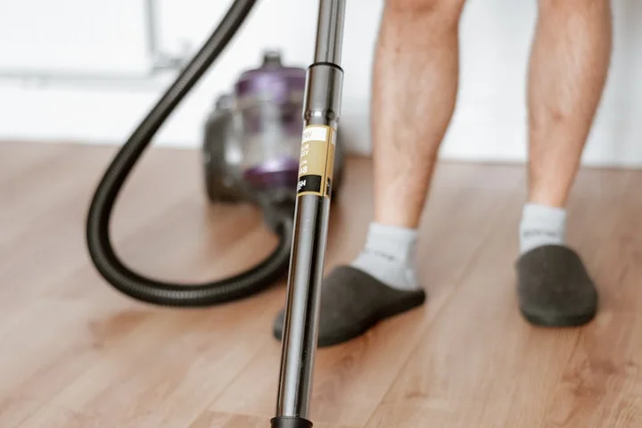Close-up of a vacuum cleaner nozzle on a wooden floor during cleanup, with a person's legs and socks visible in the background.