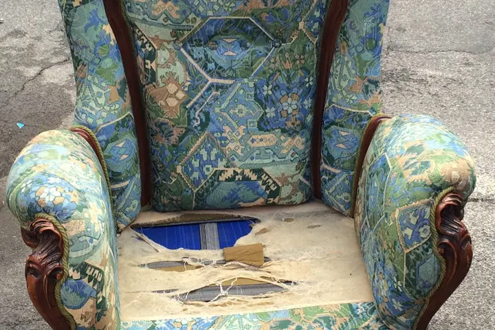 Worn antique armchair with torn patterned upholstery and exposed padding on a sidewalk