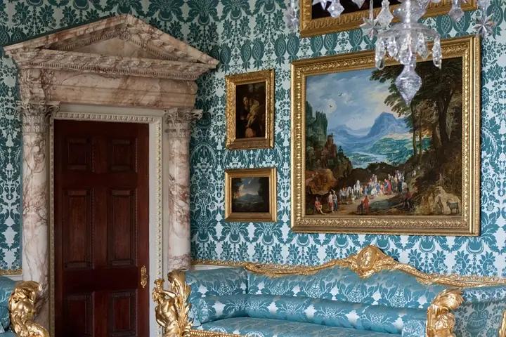 Luxurious historic parlor featuring teal damask wallpaper, a blue tufted sofa with gilded wood, and ornate framed landscape paintings.
