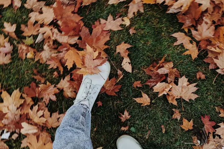 A person in light blue jeans and white sneakers walking on grass covered with orange and brown autumn leaves.