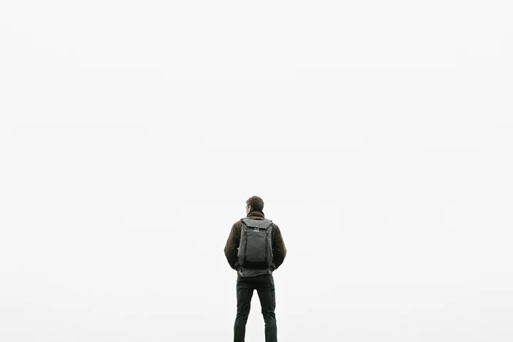 Back of a person wearing a backpack, standing alone against a bright white background.