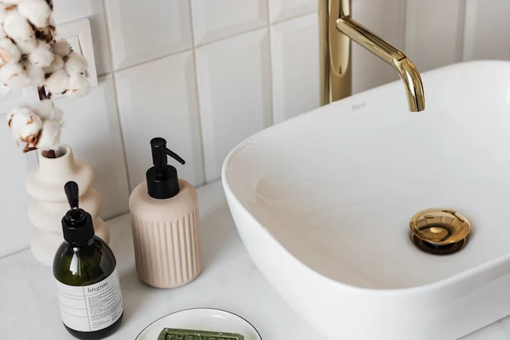 White porcelain bathroom sink with a gold faucet, cleaning bottles, and cotton stems on a marble countertop