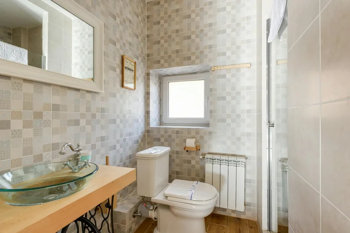 Bathroom with light mosaic tile walls, a glass vessel sink on a wooden countertop, a toilet, and a small window.
