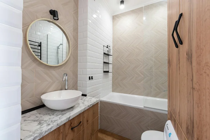 Bathroom with a marble countertop, vessel sink, round mirror, wooden vanity, and beige herringbone tile walls around a bathtub.