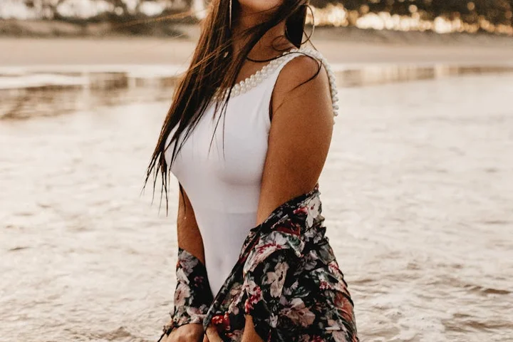 Woman by the shoreline wearing a white one-piece swimsuit and a floral cover-up, with calm water in the background.