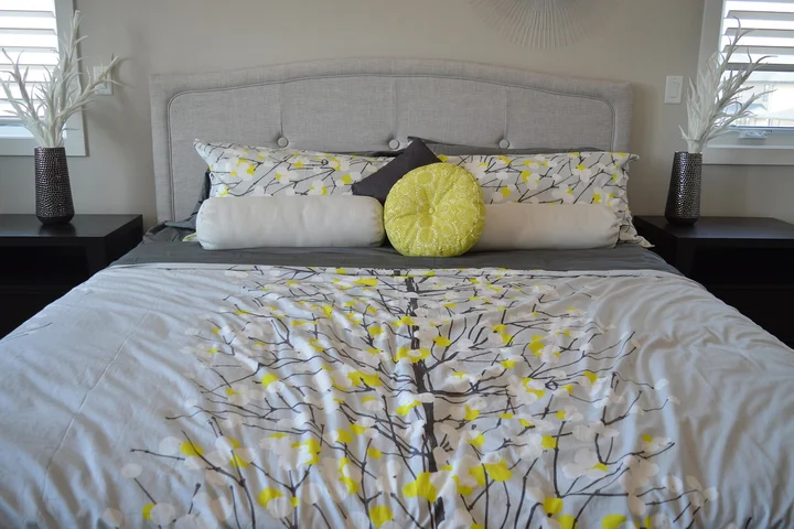 Neatly made bed with white comforter featuring gray and yellow branch patterns, two pillows, a yellow round decorative pillow, and dark nightstands with potted plants on either side.