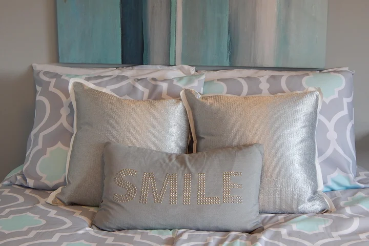 Bed with gray, white, and light blue patterned bedding and decorative pillows; a silver lumbar pillow with the word SMILE.