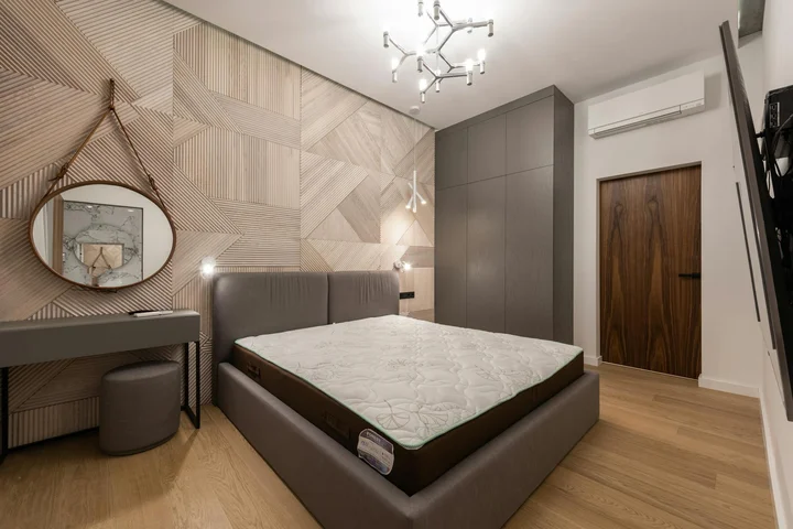 Modern bedroom with a platform bed and a bare mattress, wood floor, geometric wallpaper, a round mirror, and a tall wardrobe.