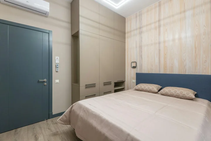 Modern bedroom with a large bed against a blue headboard, built-in wardrobe, blue door on the left, and an air conditioner mounted high on the wall.