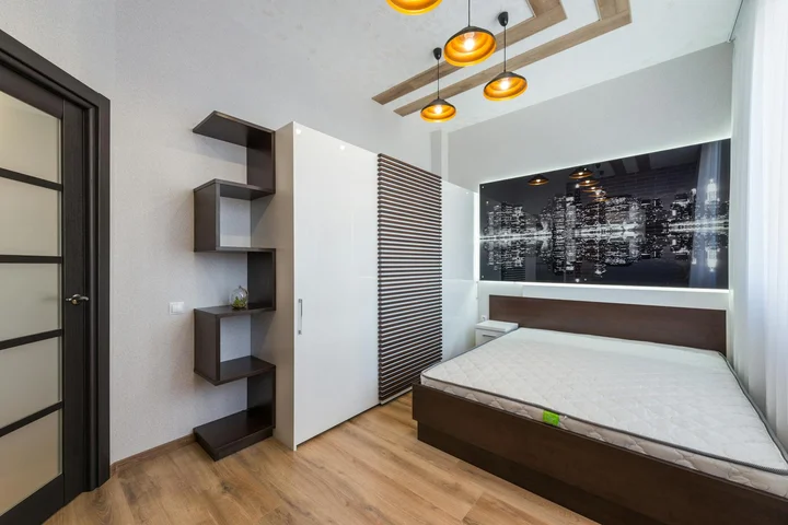 A clean, modern bedroom featuring a bed with a mattress, white wardrobe, and floating shelves, illustrating a calm environment suitable for mattress deodorizing and refresh.