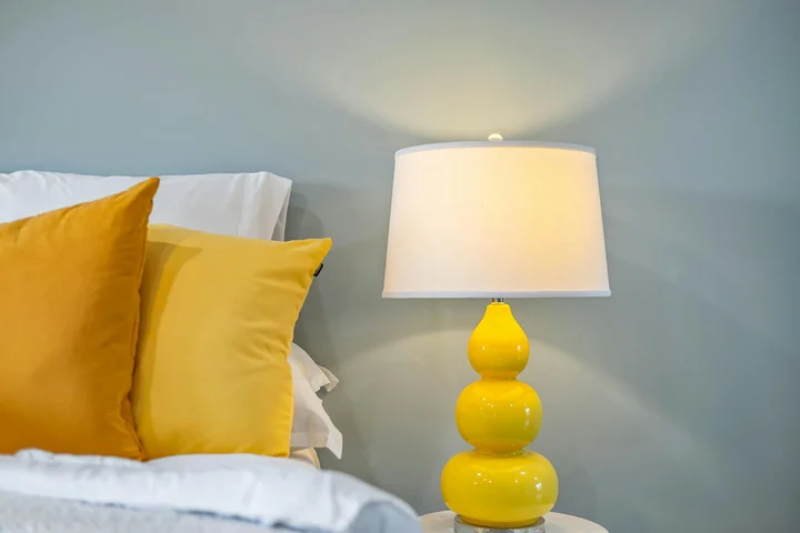 Neatly made bed with white sheets, yellow pillows, and a bright yellow lamp on a white nightstand.