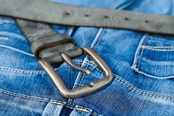 Close-up of a metal belt buckle resting on blue denim jeans, a common workwear fabric.