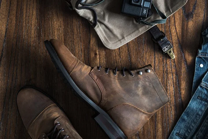 Brown leather boots with laces resting on a dark wooden floor, with clothing and a camera nearby.