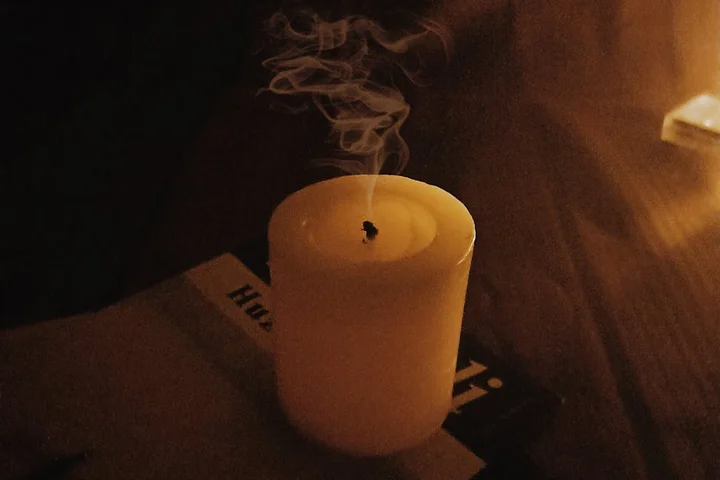 A lit white pillar candle on a dark wooden surface with wisps of smoke rising in a dimly lit room.