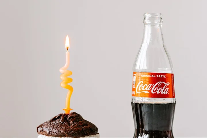 A lit candle embedded in a chocolate cupcake sits next to a glass Coca-Cola bottle on a plain light background.