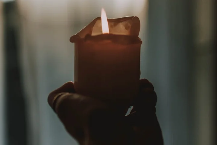 A lit candle held by a dark-gloved hand, with a warm flame and melted wax visible on the candle.