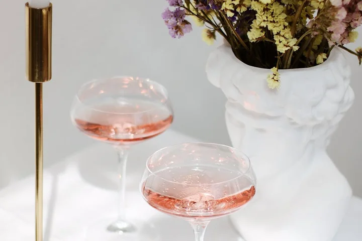 Brass candle holder and two pink-tinted glasses beside a white vase with dried flowers in a bright, minimal setting.