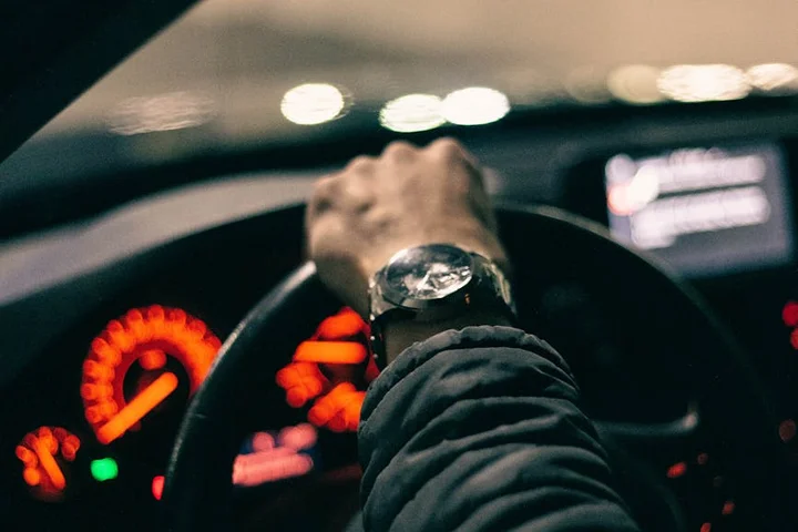 A driver's hand on the steering wheel in a dimly lit car, with the dashboard gauges glowing orange in the background.