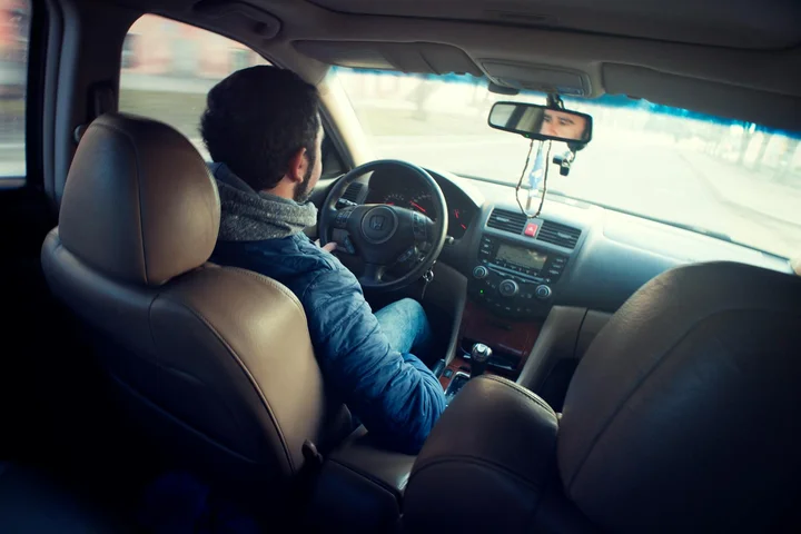 Driver in a blue jacket seated in the driver's seat of a car, with the dashboard and steering wheel in view.