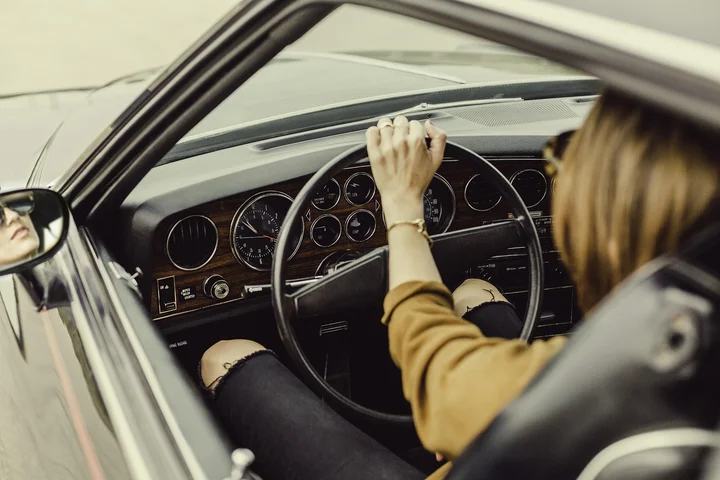 Person seated in the driver's seat of a car, gripping the steering wheel with the dashboard and upholstery visible.