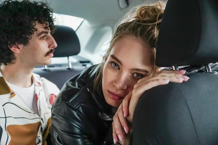 A man and a woman sit in the backseat of a car; the woman rests her head on the front seat while looking toward the camera.