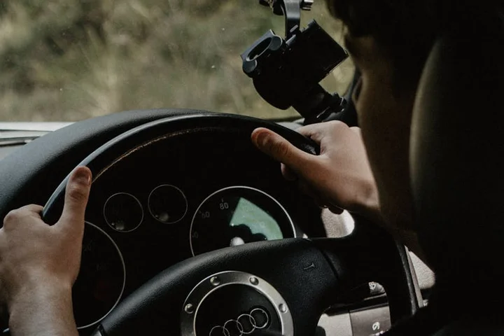 Close-up of a driver’s hands gripping the steering wheel inside a car, with the dashboard and instrument cluster visible.