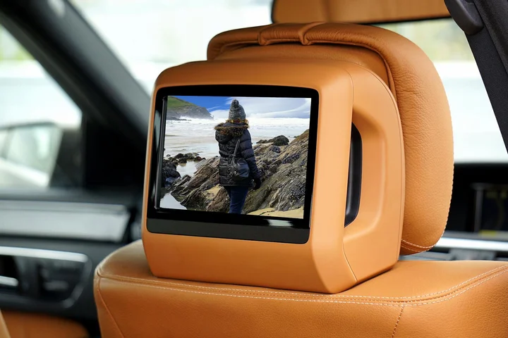 Rear passenger seat with an orange-tan headrest and a mounted screen displaying a person standing on rocks by the sea.