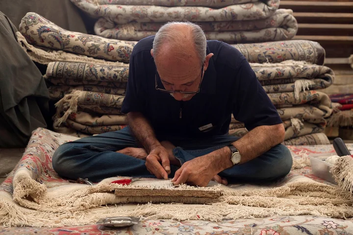 An older man sits cross-legged on a carpet, inspecting a bleach stain, with stacks of rolled carpets in the background.