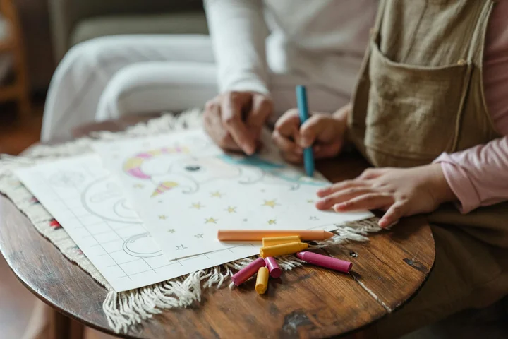 Child drawing with crayons on paper at a wooden table; art supplies like markers and crayons are nearby.