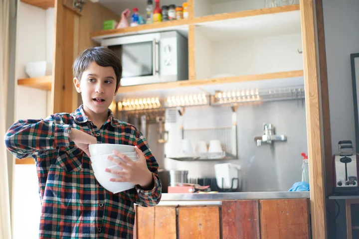 A child wearing a plaid shirt holds a white bowl in a kitchen with a microwave in the background.