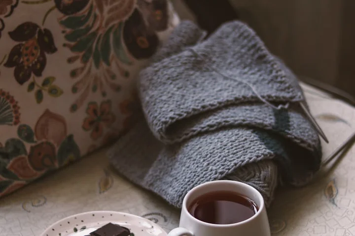 Close-up of a cozy table scene: a folded gray knit blanket, a floral-pattern bag, a small plate with a piece of dark chocolate, and a cup of tea on a light tablecloth.