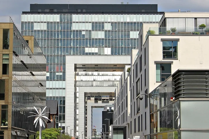 Urban cityscape with glass storefronts and a large glass-fronted building connected by a rectangular archway.