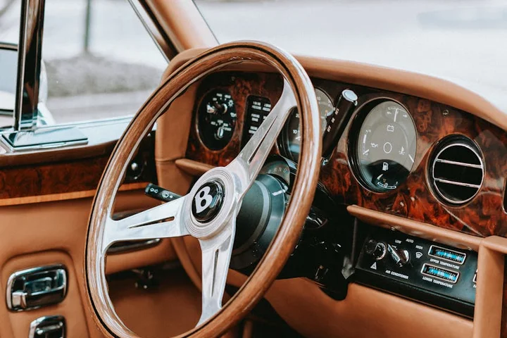 Close-up of a classic car interior with tan Nappa leather on the dashboard and seats, wooden trim, and a chrome steering wheel.