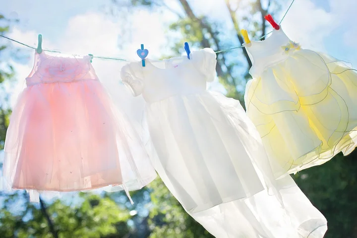 White and pastel-colored baby clothes hanging on a clothesline outdoors on a sunny day.