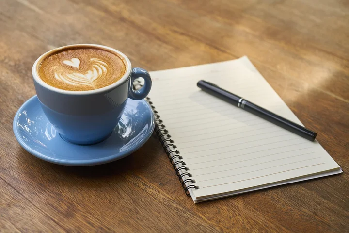 Blue ceramic cup with latte art on a matching blue saucer beside a spiral notebook and a pen on a wooden table