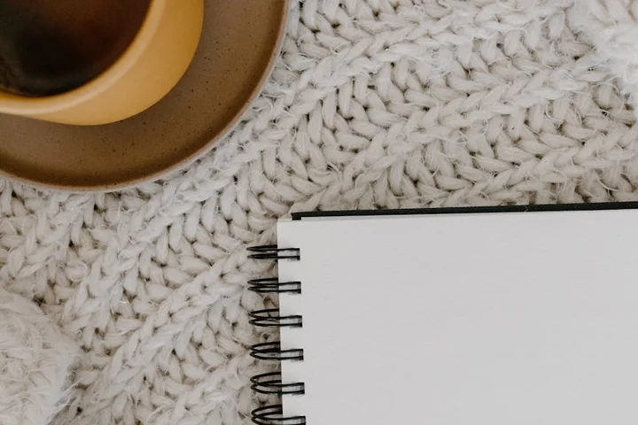 Top-down view of a cup of coffee on a white knitted blanket beside an open spiral notebook.