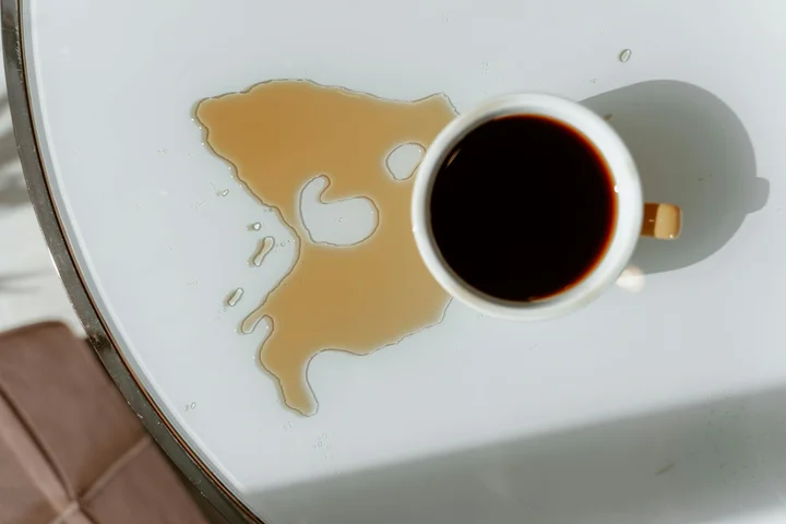 Top-down view of a coffee spill on a white surface with a cup of black coffee nearby, illustrating an oil-based stain