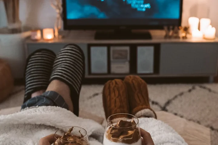 Cozy living room scene with a person sitting on a sofa wearing striped socks and slippers, holding two glasses of coffee; coffee mugs on a TV stand in the background.