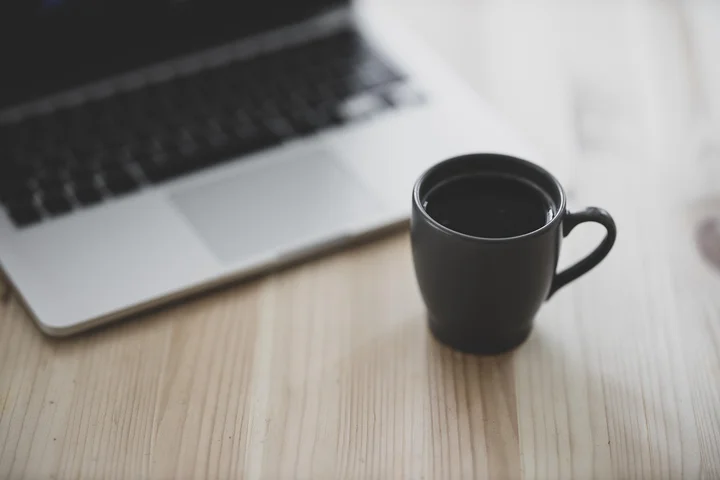 Black coffee in a mug on a light wood desk with a laptop in the background.