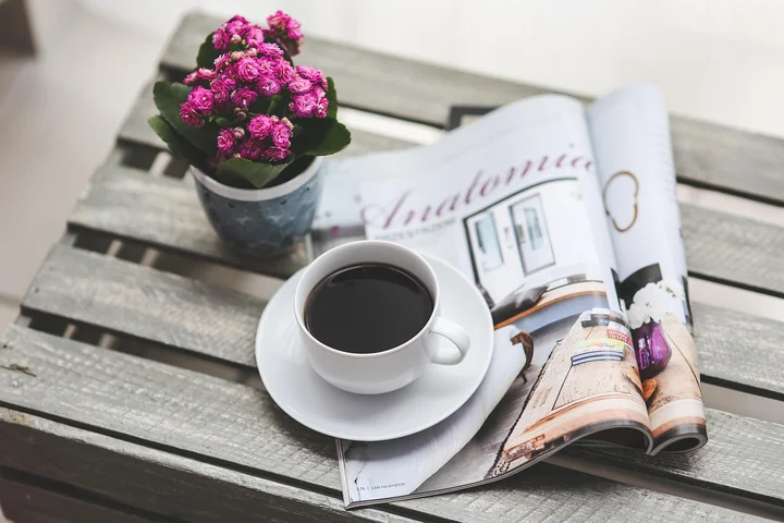 A cup of black coffee on a white saucer sits on a weathered wooden table beside an open magazine and a small potted plant with pink flowers.
