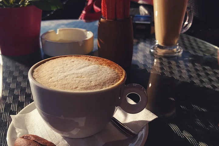 Close-up of a white ceramic coffee cup with foam on a saucer on a sunlit outdoor table.