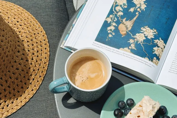A cup of coffee sits on a table beside an open magazine, with a woven straw hat on the left and a plate with blueberries and a small cake slice nearby.