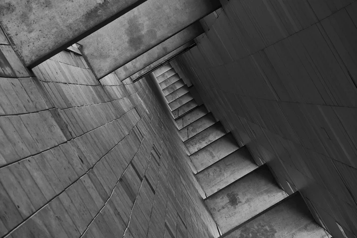 Black-and-white image of a narrow concrete stairwell viewed from the bottom, with slanted walls and repeating steps receding into the distance.