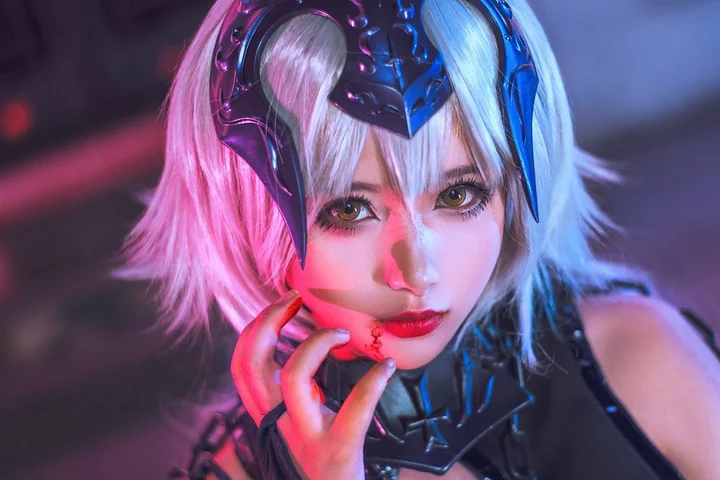 Close-up of a cosplayer with a metallic blue and pink headpiece, vivid makeup, and a finger touching their lips.
