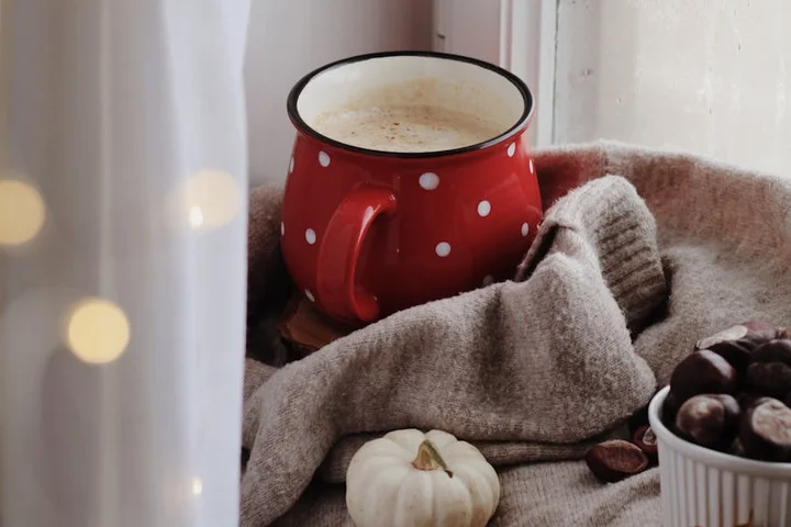 Close-up of a red polka-dot ceramic mug with coffee on a cozy beige blanket, with a small white pumpkin and a bowl of chestnuts nearby.