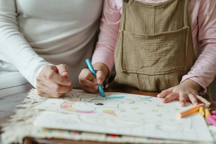 Two children drawing with crayons on a sheet of paper at a table; crayons and pencils are scattered nearby.