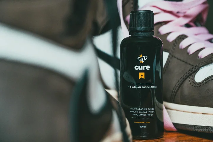 A bottle of Crep Cure shoe cleaner sits on a wooden surface with a brown and white sneaker in the background.