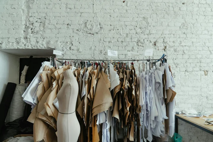 A clothing rack filled with beige, white, and gray garments in a bright studio with a white brick wall.