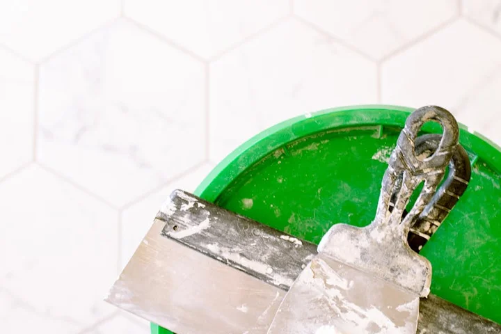 A metal putty knife resting on the rim of a green bucket, with a pale tiled wall in the background.