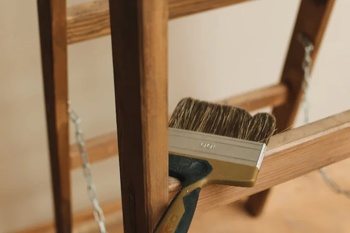 Close-up of a paintbrush applying wood stain to a wooden deck railing.